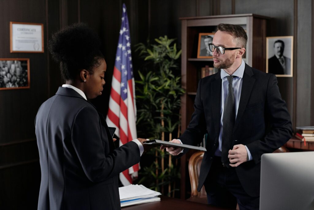 Business professionals engage in a meeting in a formal office environment with an American flag in the background.